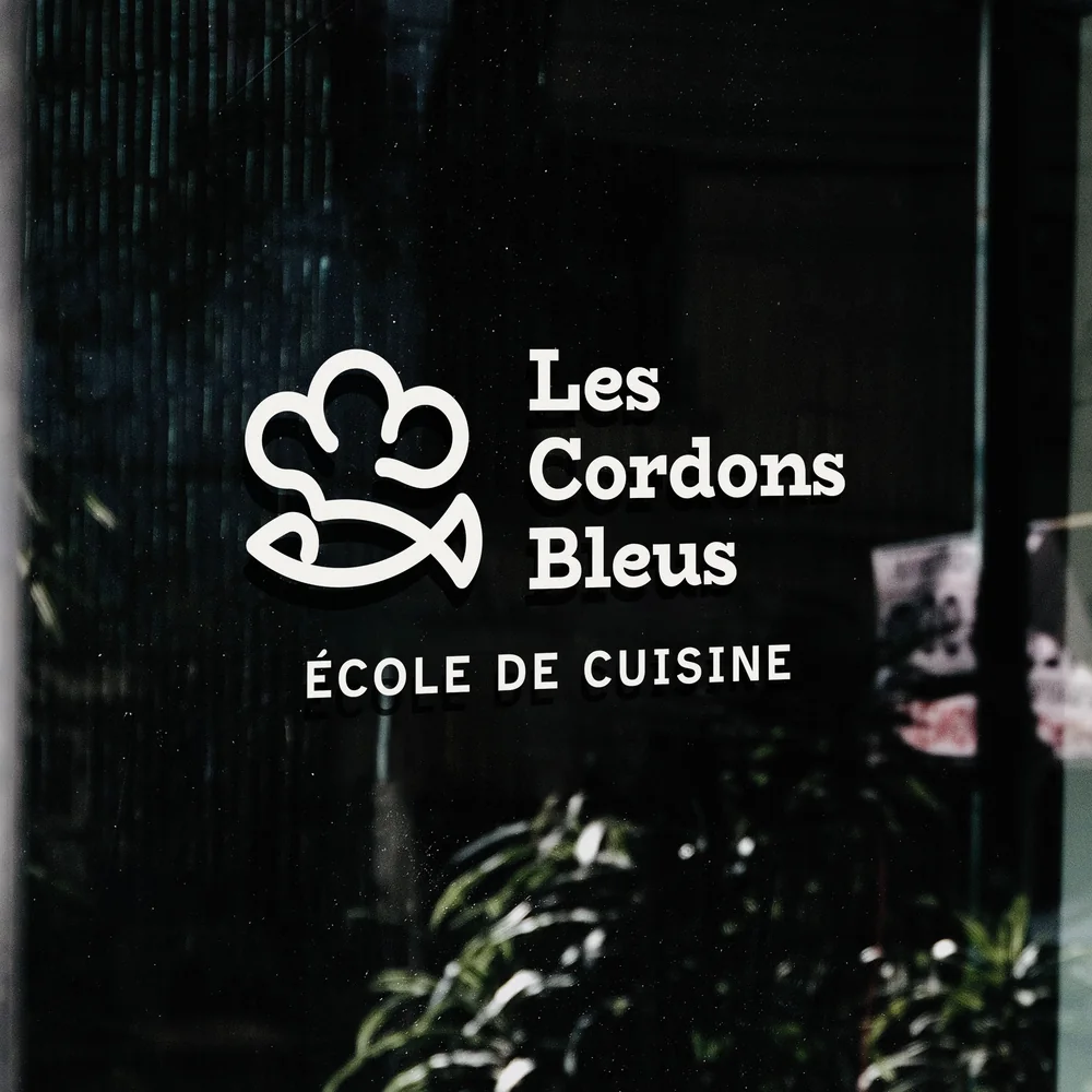 The white Les Cordons Bleus logo, “École de cuisine”, on the school's shop window, behind plants